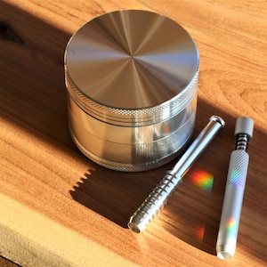 May include: A stainless steel herb grinder and two metal smoking accessories are displayed on a wooden surface. The grinder is cylindrical with a textured edge. The accessories are a metal tube and a pen-like device.