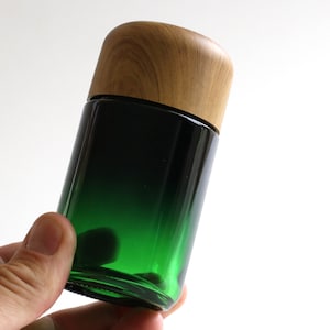 May include: A green glass bottle with a wooden lid. The bottle has a gradient from dark green to light green.