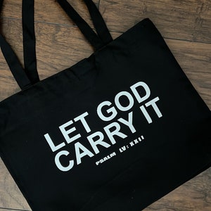 May include: Black tote bag with the text "LET GOD CARRY IT" in white, along with "PSALM LV: XXII". The bag has two black handles and is made of a sturdy material. The bag is sitting on a wooden surface.
