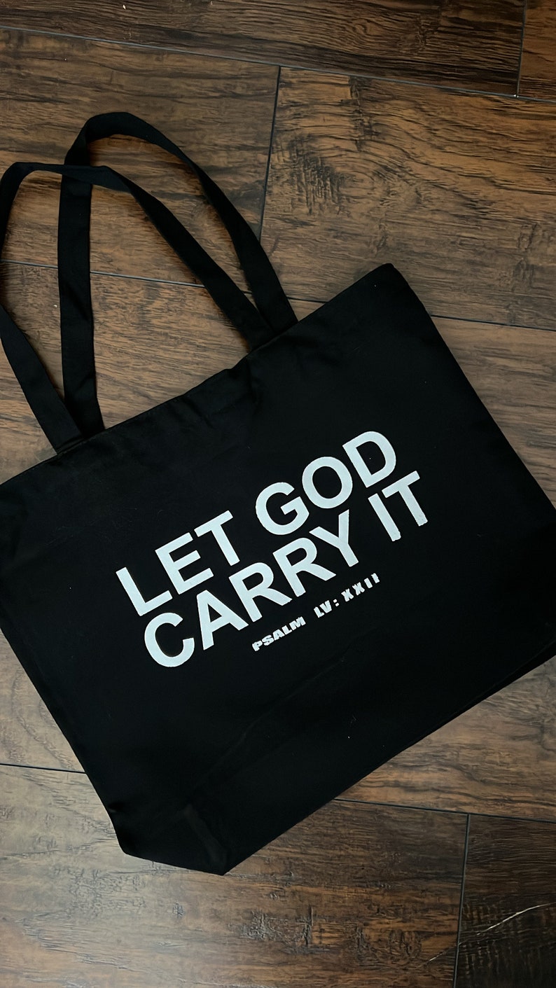 May include: Black tote bag with the text "LET GOD CARRY IT" in white, along with "PSALM LV: XXII". The bag has two black handles and is made of a sturdy material. The bag is sitting on a wooden surface.