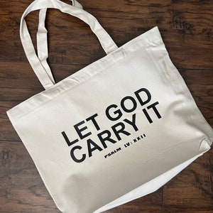 May include: Cream-colored tote bag with the text "LET GOD CARRY IT" in bold black letters. The phrase "PSALM LV: XXII" is printed below. The bag has two cream-colored handles and is made of a sturdy fabric.