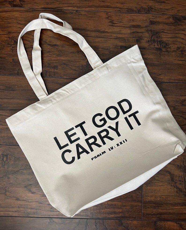 May include: Cream-colored tote bag with the text "LET GOD CARRY IT" in bold black letters. The phrase "PSALM LV: XXII" is printed below. The bag has two cream-colored handles and is made of a sturdy fabric.