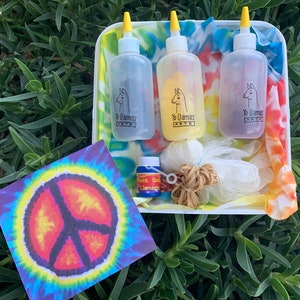 Puede incluir: Un kit de teñido anudado con tres botellas de tinte para tela, gomas elásticas, guantes y un patrón de teñido anudado con un signo de paz. Las botellas están etiquetadas como "Yo Llamas Arts" y son de color azul, amarillo y morado.