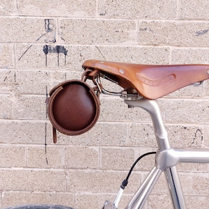 May include: A brown leather bicycle saddle with a matching cylindrical leather tool bag attached underneath. The saddle features a silver metal seat post and a stamped logo on the side. The tool bag is secured with leather straps and buckles.