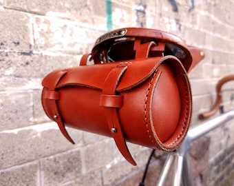 May include: A cylindrical brown leather bicycle saddle bag featuring two vertical buckle straps on the front flap. The bag is attached to a brown leather bicycle seat with a visible metal plate embossed with the brand name Brooks.