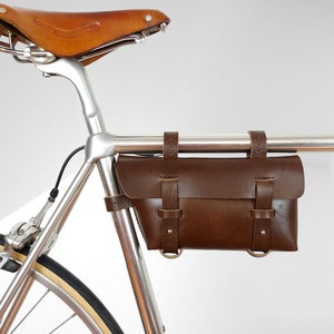 Könnte beinhalten: Eine braune Ledertasche für den Fahrradlenker mit Schnallen und Riemen, die an einem silbernen Fahrradrahmen befestigt ist.