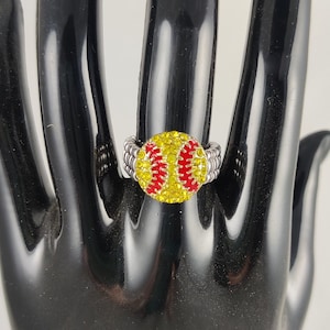 May include: A silver ring with a red and yellow baseball design. The baseball is covered in rhinestones.