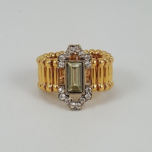 May include: A gold ring with a rectangular green stone surrounded by clear rhinestones. The ring is made of multiple bands of gold.