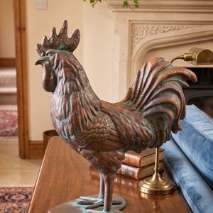 May include: A copper-toned metal rooster sculpture featuring a weathered finish with green patina accents. The rooster stands on a heart-shaped base, displaying detailed feather textures, a prominent comb, and a wattle, captured in a side profile.