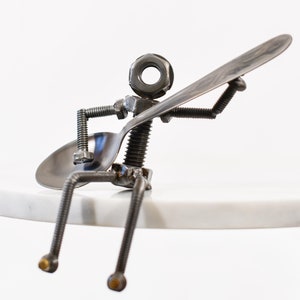 May include: A quirky sculpture of a figure made from metal components, including a spoon, bolts, and a nut for the head. The figure appears to be playing a spoon like a guitar, with the spoon held up. The sculpture is sitting on a white surface.