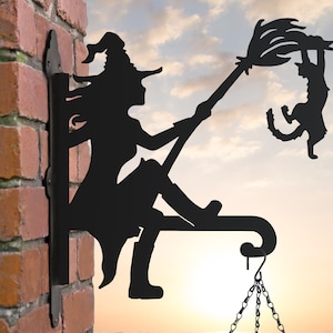 May include: Black metal silhouette of a witch riding a broomstick with a black cat hanging on. The witch is wearing a pointed hat and has long hair. The silhouette is mounted on a brick wall with a chain hanging from the bottom.