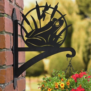 May include: Black metal wall-mounted plant hanger featuring a frog and dragonfly design. The hanger has a hook for suspending a plant pot.