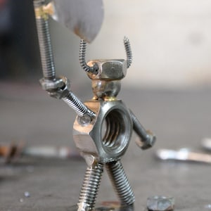 May include: A metal sculpture of a warrior figure, made from nuts, bolts, and screws. The figure holds a small axe and has curved metal pieces resembling horns. The sculpture is on a metal surface.