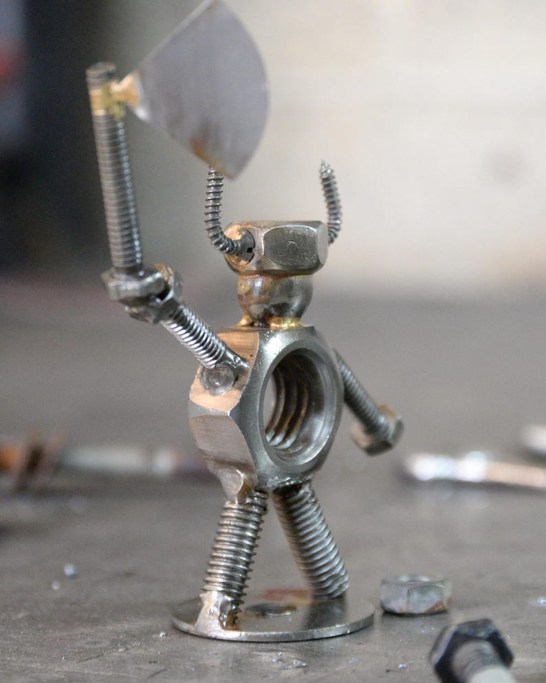 May include: A metal sculpture of a warrior figure, constructed from nuts, bolts, and screws. The figure holds a small axe and has coiled wire horns. The sculpture is silver and gray, with a round base.
