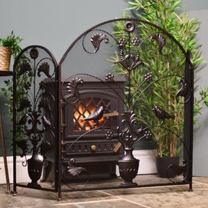 May include: A black metal fireplace screen with decorative bird and floral motifs. The screen has a mesh centre and an arched top. The fireplace behind the screen has a glowing fire, creating a warm ambiance.