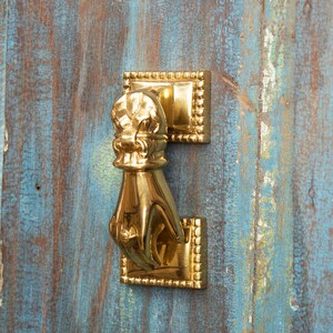 May include: A gold-toned door knocker shaped like a hand with a decorative crown on top. The knocker is mounted on a wooden door.