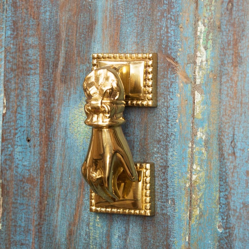 May include: A gold-toned door knocker shaped like a hand with a decorative crown on top. The knocker is mounted on a wooden door.
