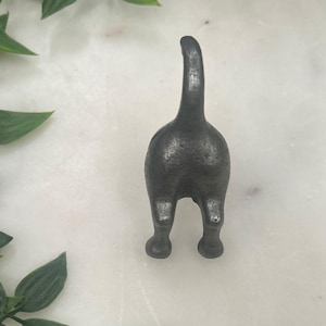 May include: A dark grey metal cat's rear figurine with a textured surface. The cat's tail is raised, and its hind legs are visible. A whimsical decorative item.
