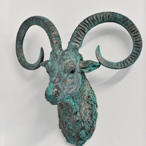May include: A green and brown resin sculpture of a ram's head with curled horns. The sculpture is mounted on a white wall.