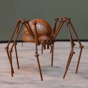 May include: A rustic metal spider sculpture with a weathered, rusty finish. The spider features a rounded body, multiple legs, and detailed eyes, creating a unique decorative piece. The sculpture is designed to stand on a surface.
