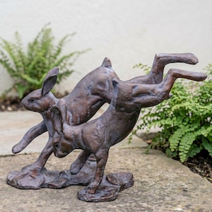 May include: A bronze-toned sculpture depicting two hares in motion. One hare is positioned in a leaping posture with its hind legs extended, while the other hare is shown in a lower, running stance. Both figures feature textured surfaces.