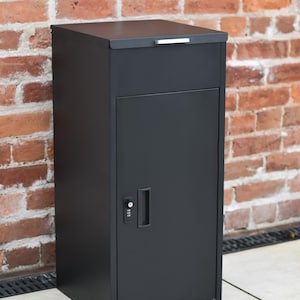 May include: A black metal mailbox with a combination lock and a hinged door. The mailbox is standing on a concrete surface in front of a brick wall.