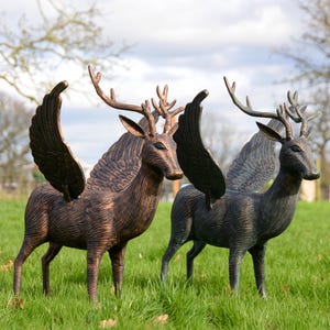 May include: Two bronze deer sculptures with large wings stand in a grassy field. The deer have detailed antlers and are posed naturally. The sculptures are a decorative garden feature, with a weathered finish.