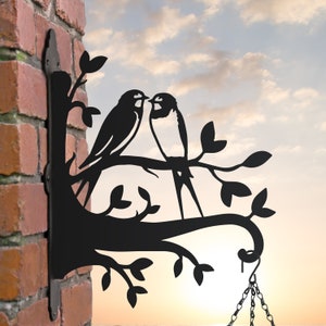 May include: Black metal silhouette of two birds perched on a branch with a decorative scroll at the end. The birds are facing each other and appear to be kissing. The branch has leaves and is attached to a wall.