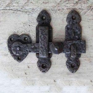 May include: A black metal heart-shaped latch with a sliding bolt. The latch has a rustic, aged finish and is designed for use on doors or gates.