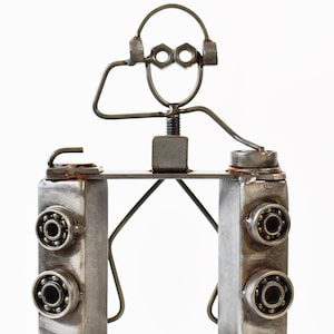 May include: A metal sculpture of a DJ figure. The figure is made from various metal parts, including nuts, bolts, and bearings. The DJ is wearing headphones and has arms outstretched towards a mixing console.