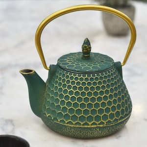 May include: A green cast iron teapot with a gold handle and lid. The teapot has a honeycomb pattern and a gold finial on the lid.