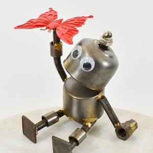 May include: A handmade metal robot sculpture holding a red butterfly. The robot features googly eyes, a silver head, and a cylindrical body. Its arms and legs are constructed from springs and bolts, creating a distinctive, industrial appearance.