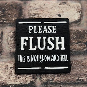 May include: A square black metal sign with white raised lettering that reads PLEASE FLUSH THIS IS NOT SHOW AND TELL. The sign features horizontal white lines above and below the text and two small circular holes for mounting on the left and right.