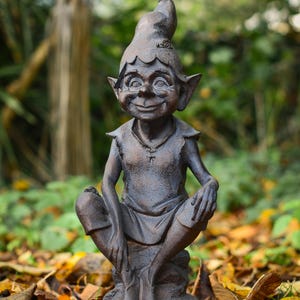 May include: A bronze garden gnome statue with a pointed hat and a smiling face. The gnome is seated with its legs crossed. The statue is surrounded by fallen leaves. A decorative garden ornament.