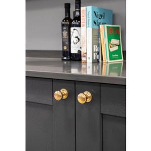 May include: Two gold cabinet knobs with a textured finish are mounted on dark grey cabinet doors. The knobs are round and have a raised design.