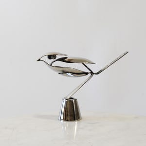 May include: A silver metal bird sculpture. The bird is crafted from spoons and other metal pieces, with a detailed design. The sculpture is mounted on a small, silver metal base. The bird's tail is made of a thin, flat piece of metal.
