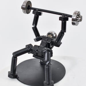 May include: A black metal sculpture of a person lifting weights. The sculpture is made of nuts and bolts and is on a black metal base.