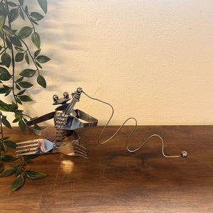 May include: A quirky sculpture made from recycled cutlery, with forks and spoons arranged to look like a musician playing an instrument. The sculpture is made of silver-coloured metal and sits on a dark wooden surface.