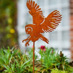 May include: A weathered, orange metal owl garden ornament, depicted in flight, with detailed wing cutouts. The owl is mounted on a stake, positioned above green plants in a metal container. The background features a window and brickwork.