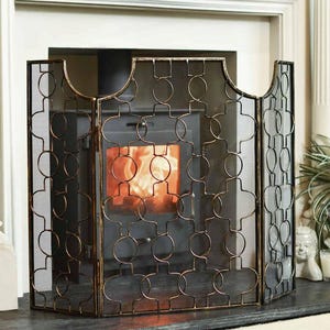 May include: A three-panel fireplace screen featuring a black metal frame with a gold-toned distressed finish. The screen has a repeating geometric pattern of interlocking circles and vertical bars, backed by a fine black mesh metal screen.