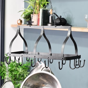 May include: A black metal pot rack hanging from chains. The rack has a rectangular shelf, curved supports, and hooks for pots and pans. Kitchen storage solution.