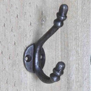 May include: A dark grey, cast iron coat hook with a decorative design. The hook is mounted on a light-coloured wooden surface. The hook has a rounded base with two screw holes and a curved arm with decorative finials.
