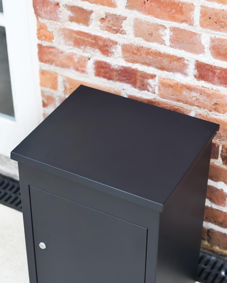 May include: A black, rectangular postbox with a door and a small silver knob. The postbox is set against a red brick wall and a white door frame.