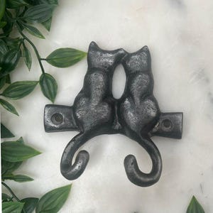 May include: A dark grey metal wall hook featuring two cat silhouettes facing each other. The hook has a horizontal bar with mounting holes and curved tails for hanging items. The design is a decorative accent for a home.