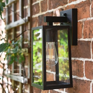 May include: A black, wall-mounted outdoor lantern with a rectangular glass enclosure. It features a vintage-style light bulb and is mounted on a red brick wall. The design is modern and suitable for exterior lighting.