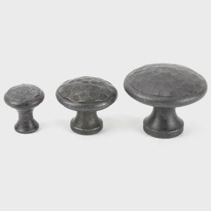 May include: Three dark grey cabinet knobs with a hammered texture. The knobs are mushroom-shaped, with a rounded top and a short, cylindrical base. The sizes vary, with the largest knob on the right and the smallest on the left.