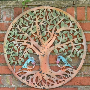 May include: A circular metal wall art piece featuring a tree with intricate branches and roots. The tree has light green leaves and a copper-toned trunk. Two small blue birds with pink chests are perched on the roots at the base.