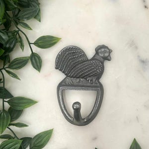May include: A cast iron rooster-shaped wall hook in a dark grey finish. The hook features a detailed rooster design atop a curved hook, ideal for hanging items. The hook is mounted on a white marble surface with green leafy accents.