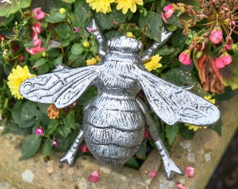 Puede incluir: Adorno de jardín de metal plateado con forma de abeja, con alas y cuerpo detallados, colocado entre flores amarillas y rosas y follaje verde. La abeja es un elemento decorativo.