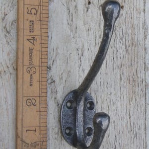 May include: A dark grey, cast iron coat hook with a curved design. The hook has a rounded top and a lower hook, with an oval base featuring four screw holes. A wooden ruler is next to the hook.